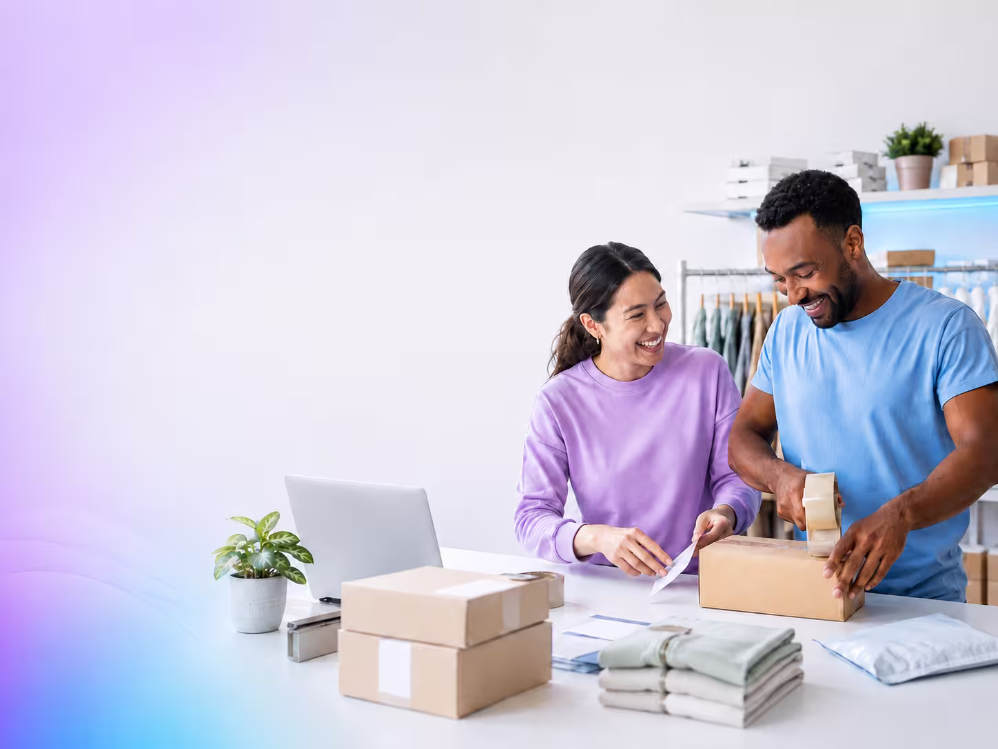 Two people packing customer orders at a workbench in a small retail studio, laptop open beside them